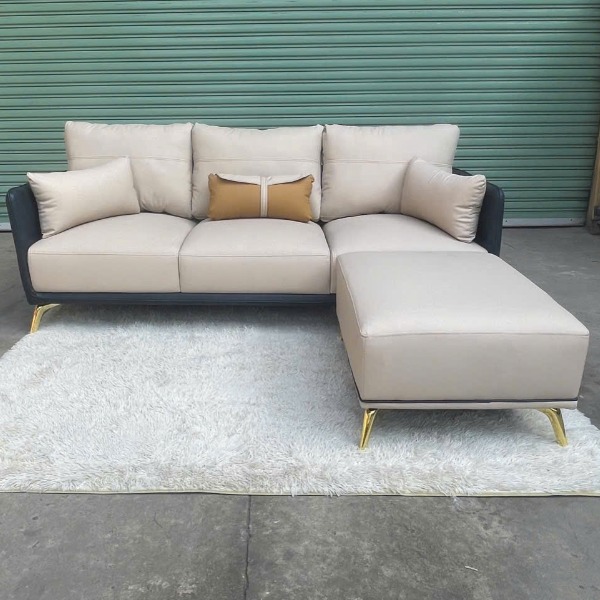 Bộ sofa góc chữ L da màu kem, chân inox mạ vàng sang trọng, đi kèm gối tựa và đôn.